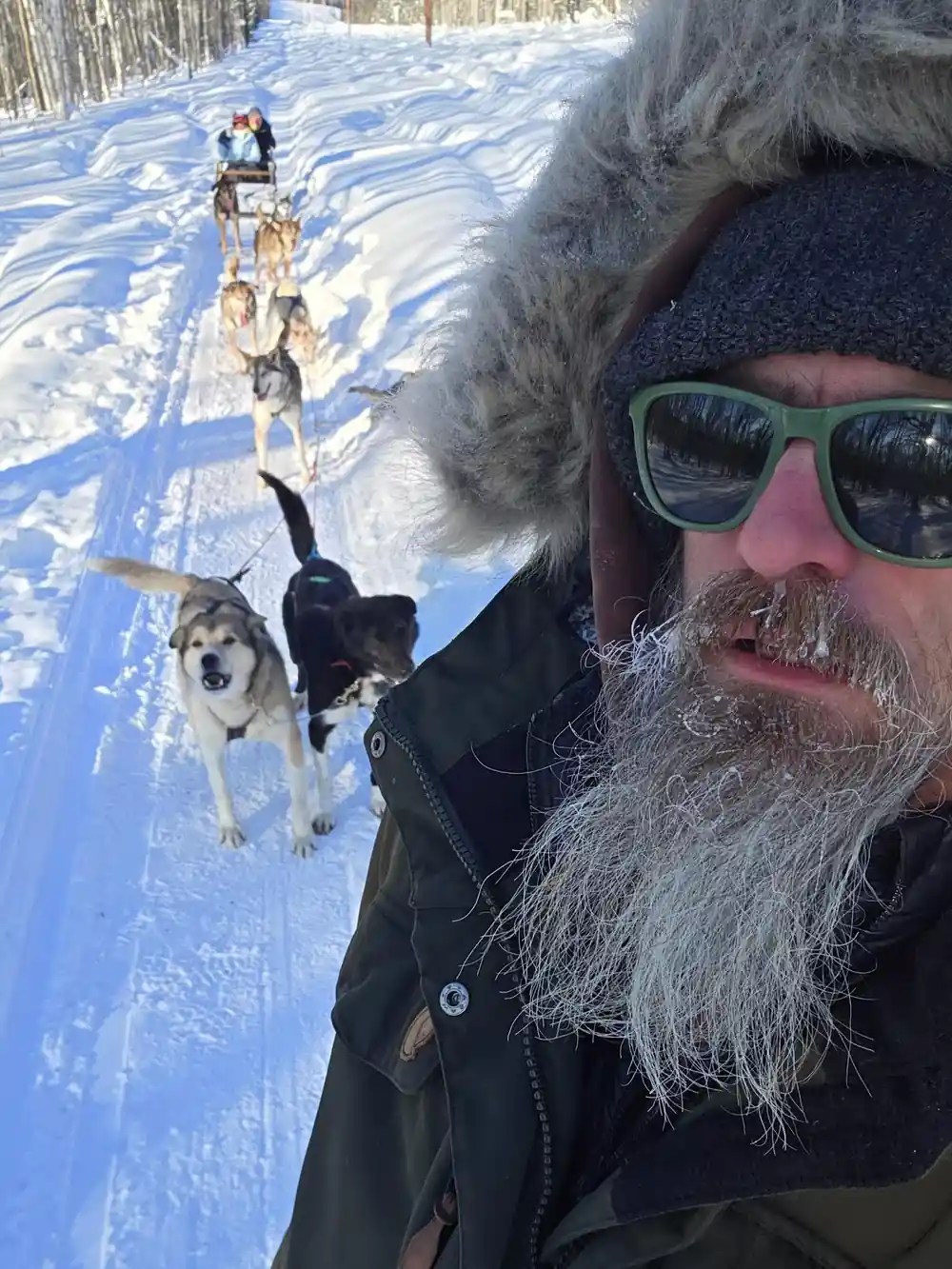 Musher selfie with a frosted beard, dogs on the trail behind