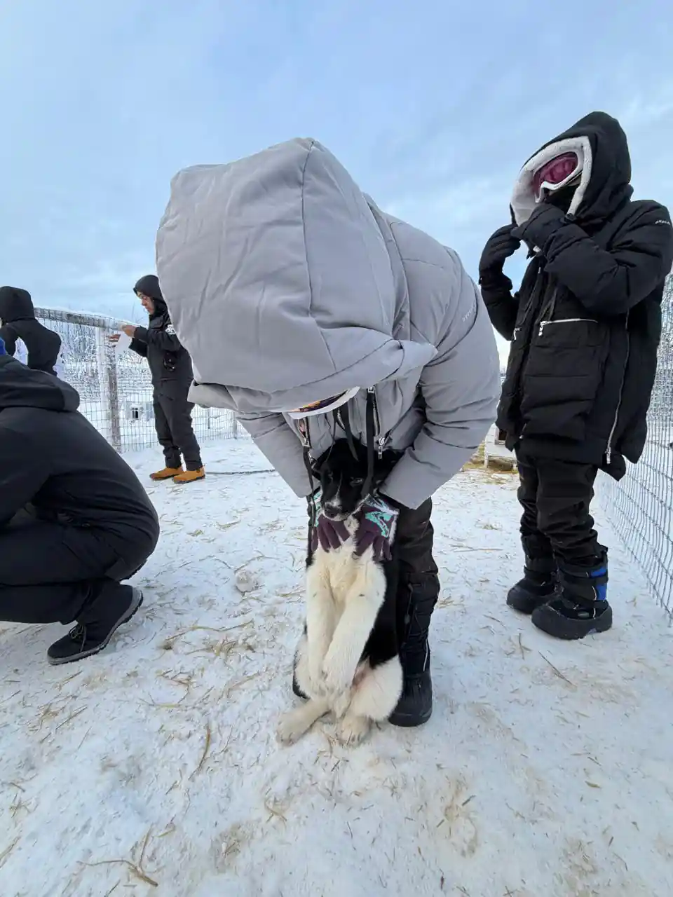 Visitors in heavy hooded parkas beside a dog at the kennel
